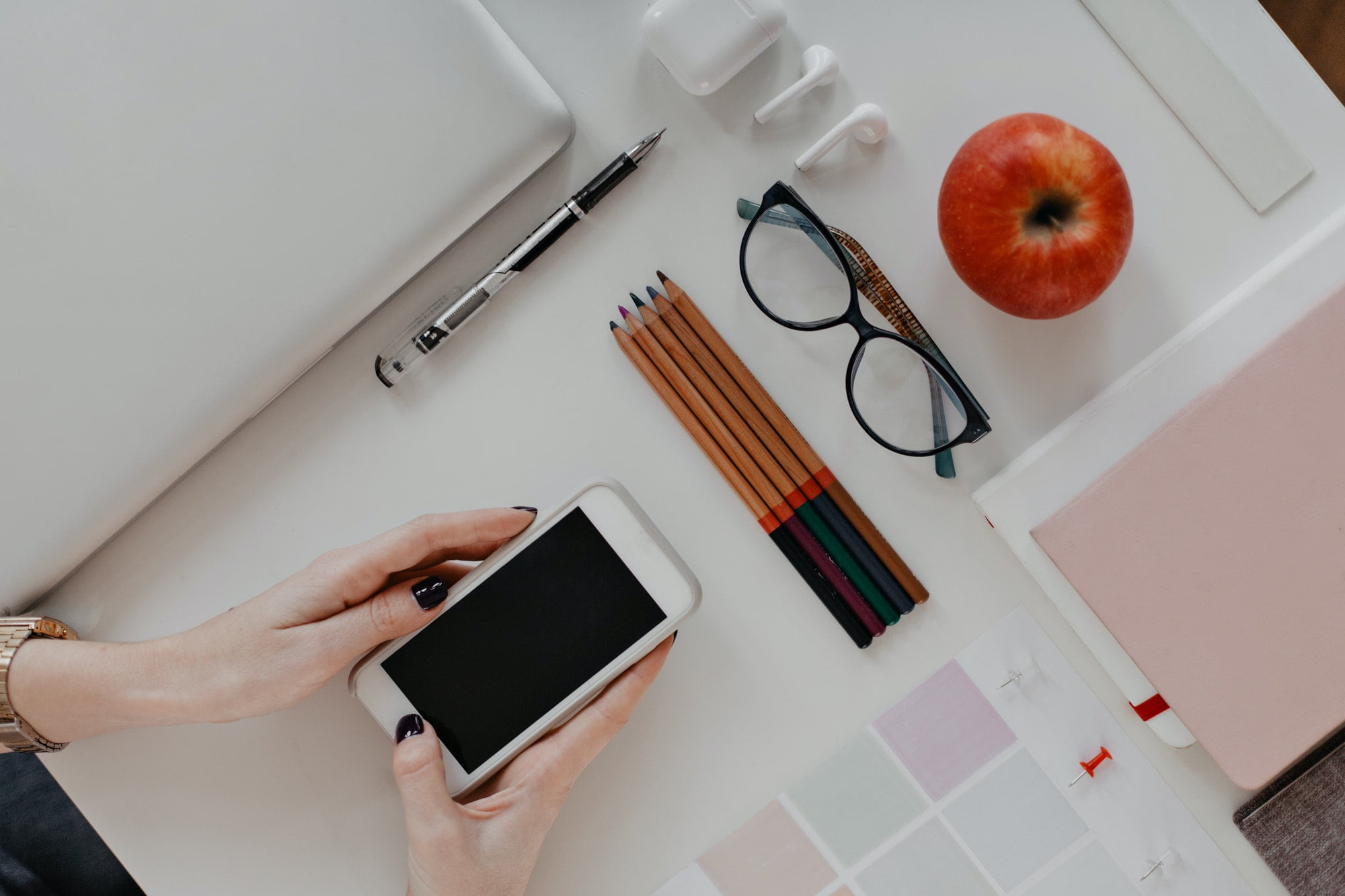 Flat lay. Womans hands holding iPhone on background of pencils, glasses, MacBook