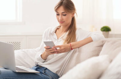 Woman Texting On Phone Working On Laptop At Home