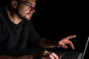 A man with glasses works at a laptop in the dark.