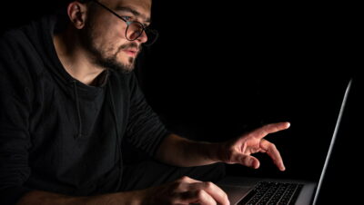 A man with glasses works at a laptop in the dark.