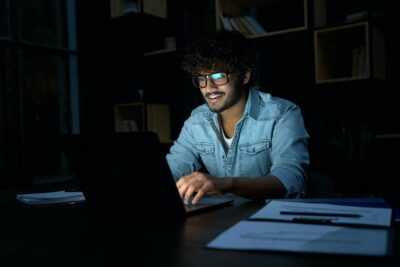 Smiling young indian business man working online late at night using laptop.