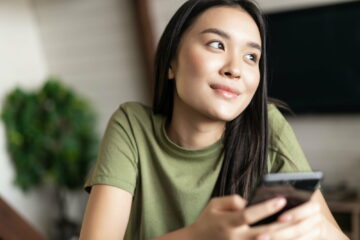 Young dreamy woman looking outside window, holding smartphone and smiling, chatting on phone app