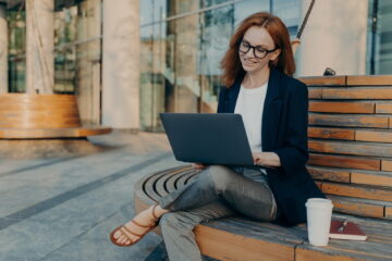 Intelligent redhead lady browses laptop outdoors makes project online