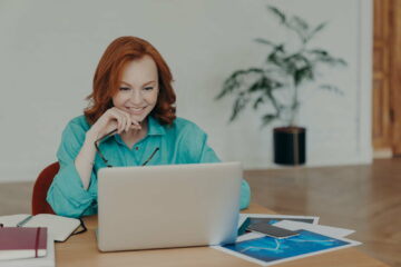 Woman with red hair works or studies on modern laptop computer, browses internet.