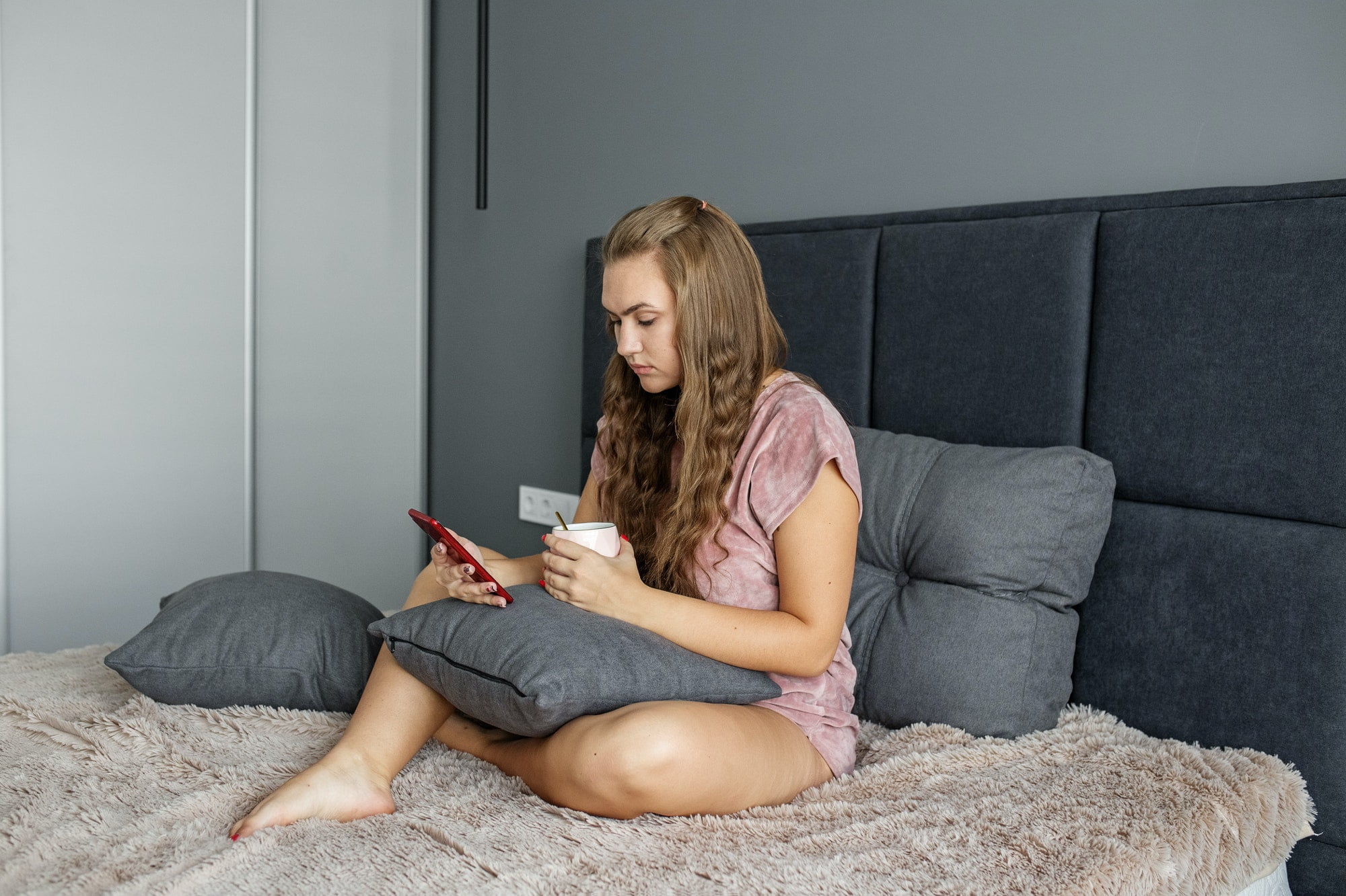 A teenage girl sits in bed and drinks tea. Uses mobile phone. A woman is texting on social networks