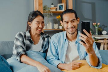 man and woman sit couch use smartphone facetime video call with friends and family in living room.