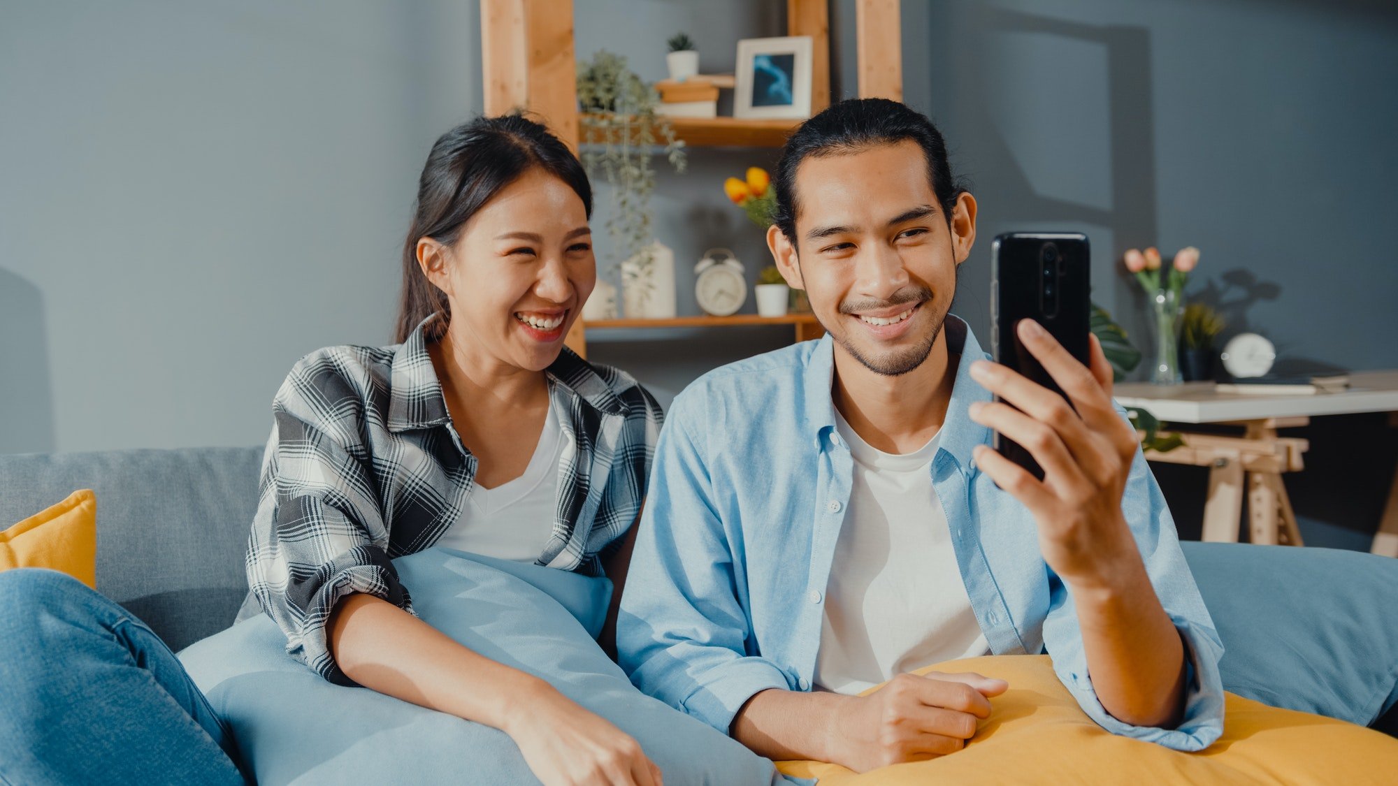 man and woman sit couch use smartphone facetime video call with friends and family in living room.