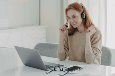 Pleasant smiling redhead woman in headset communicating with colleagues during video call on laptop