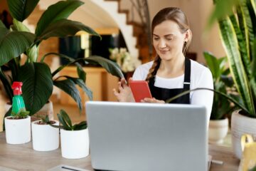 A young female florist checks orders in the phone app and on a laptop, takes orders online.