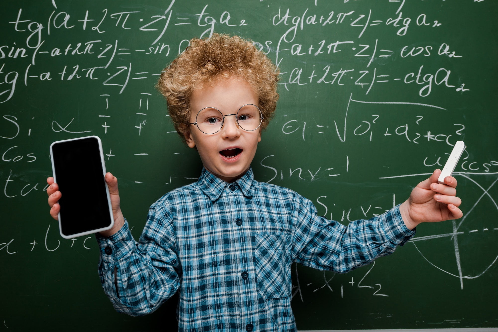 cute and smart child holding smartphone with blank screen and holding chalk near chalkboard with