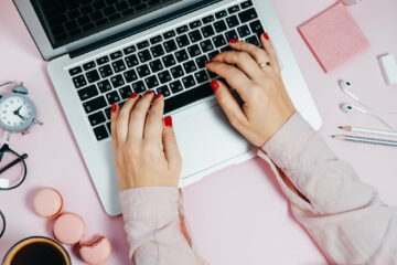 Female hand writing on Laptop on pink plant macaroons alarm clock Top View