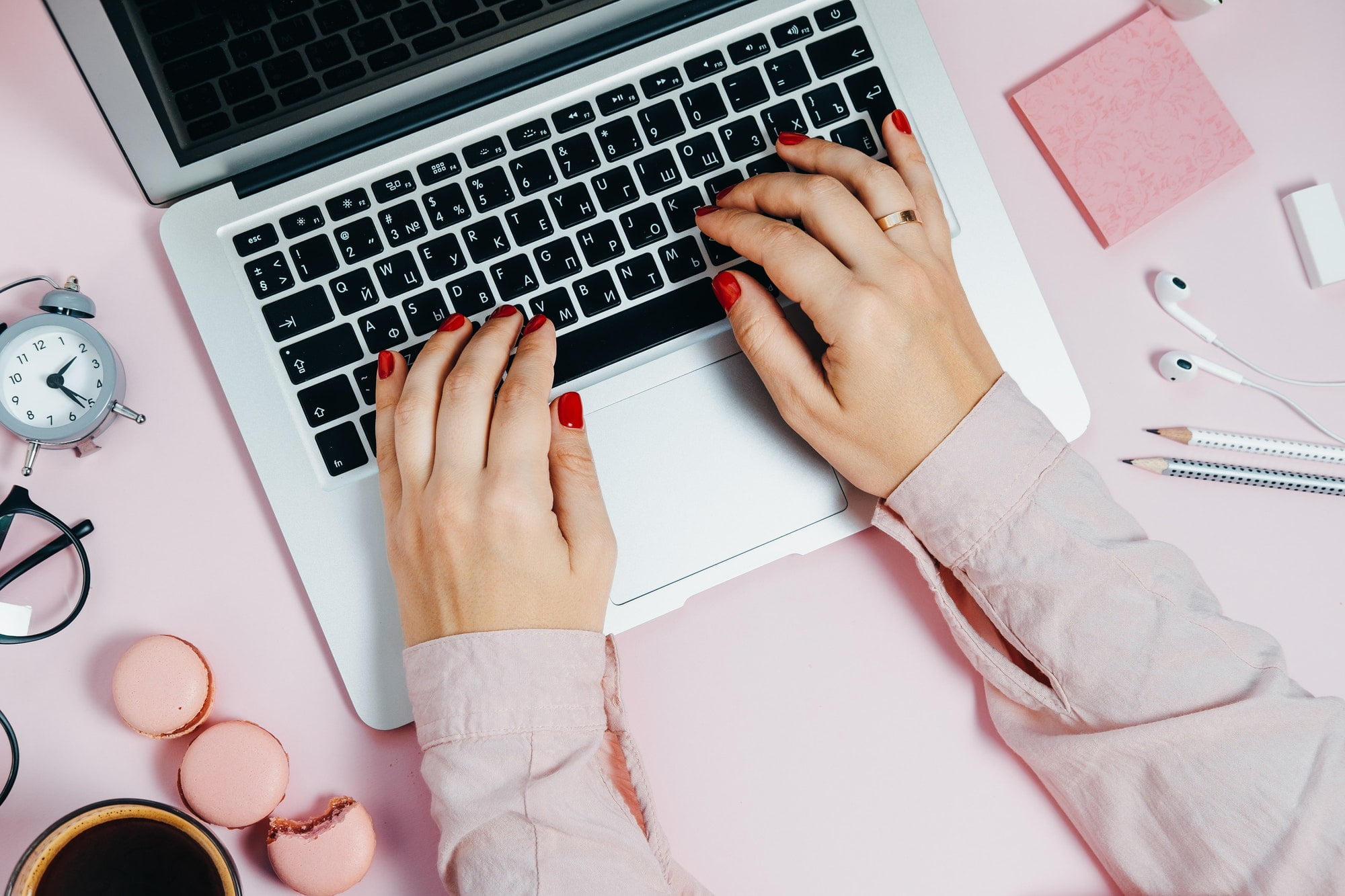 Female hand writing on Laptop on pink plant macaroons alarm clock Top View