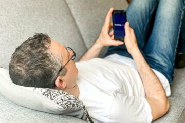 Middle aged adult man is doing booking hotel at Booking.com app at home on sofa using mobile phone