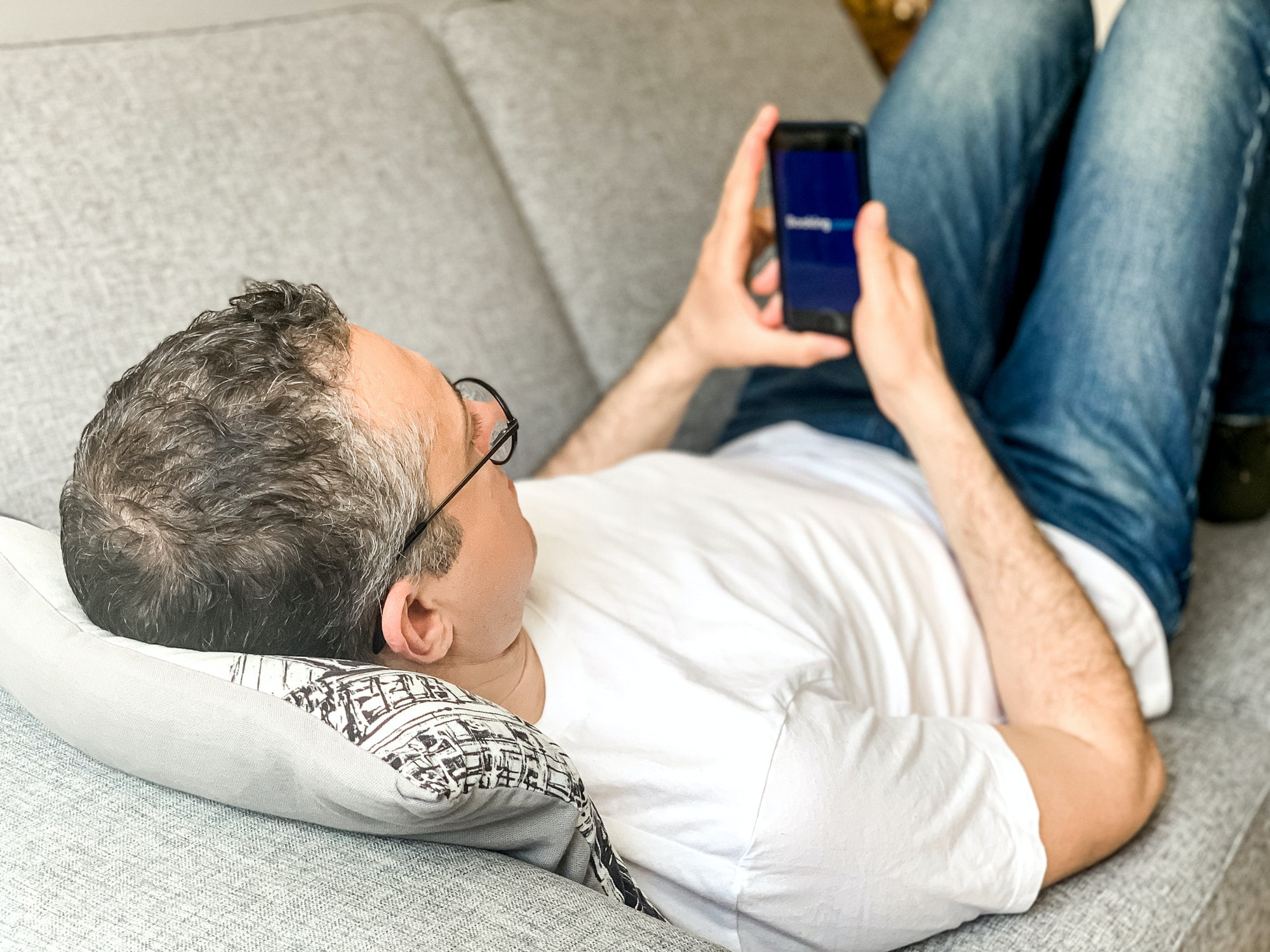 Middle aged adult man is doing booking hotel at Booking.com app at home on sofa using mobile phone