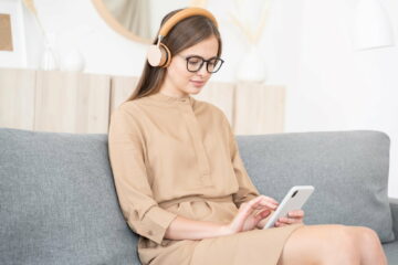 Young female sitting on gray sofa with phone, typing messages, listening to music