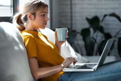 Confident woman working with her laptop while drinking a cup of coffee sitting on a couch at home