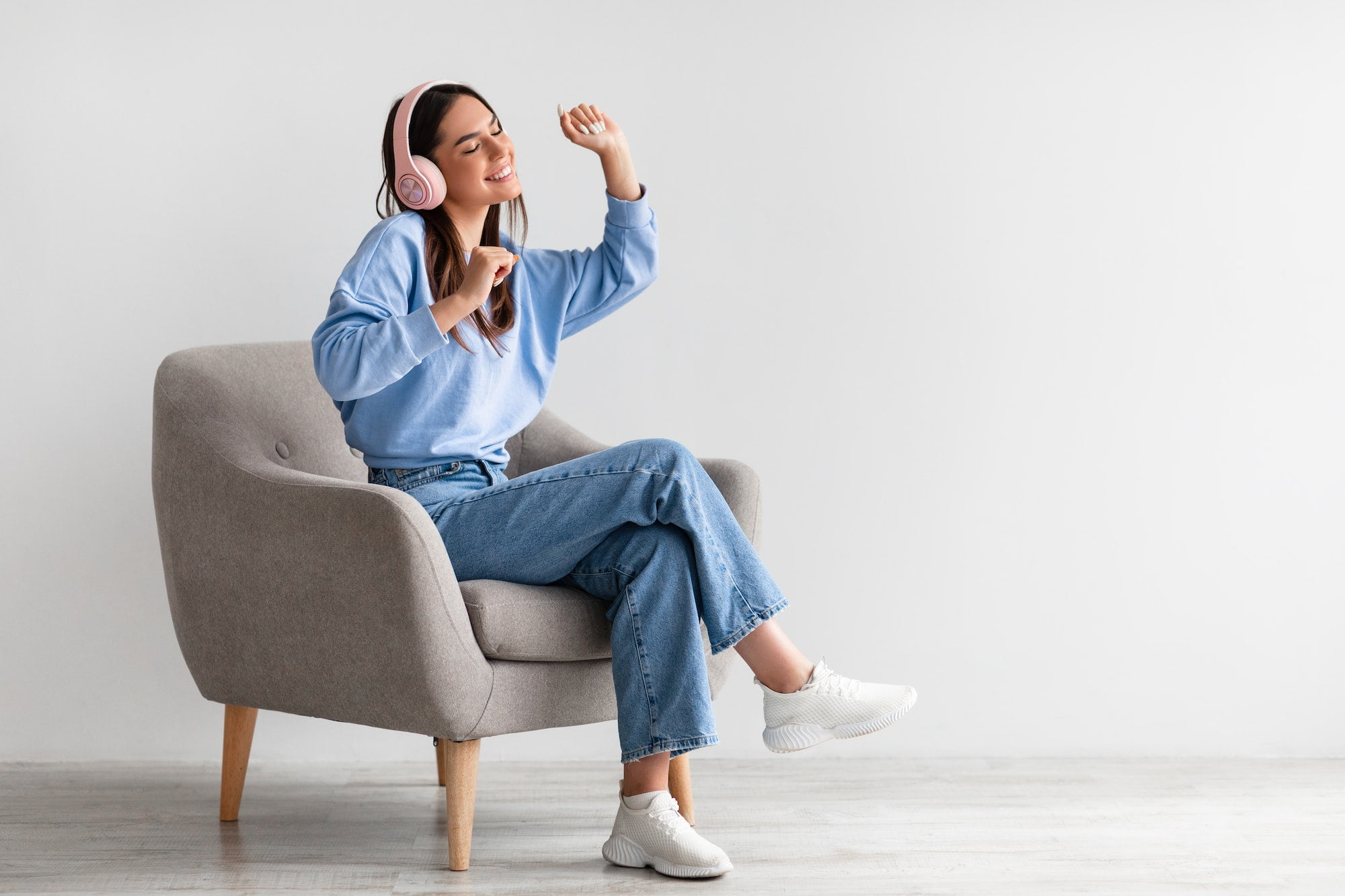 Happy millennial lady in wireless headphones relaxing in armchair and enjoying music against white