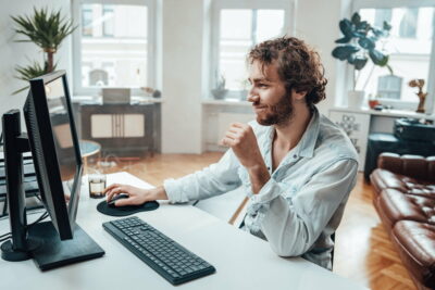 Joyful guy with curly hairs and beard sits at table working on pc