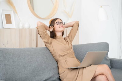 Young lady sitting on couch with open laptop, listening to music via wireless headphones