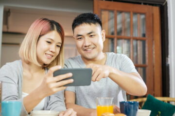 asian couple in pajamas sitting at table iand watching video on mobile having breakfast