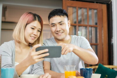 asian couple in pajamas sitting at table iand watching video on mobile having breakfast