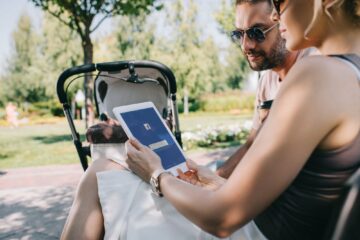 parents sitting near baby carriage in park and looking at tablet with loaded facebook page