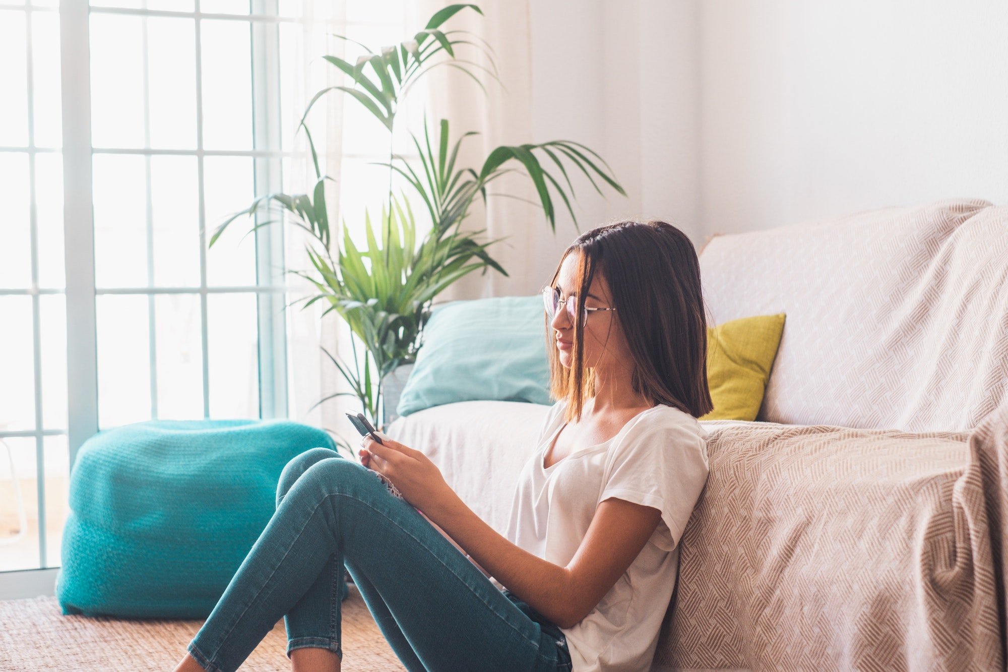 Serious woman in eyeglasses text messaging using mobile phone while sitting on floor