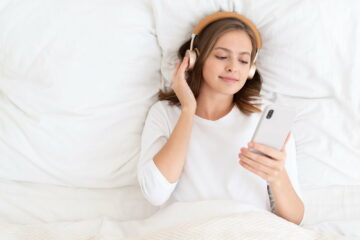 Young girl lying on white bed in the morning, holding smartphone and listen to favorite music