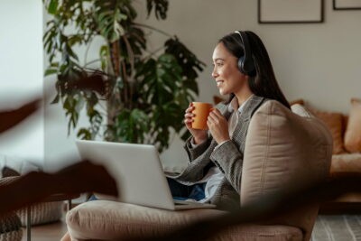 Attractive asian business woman listening music on headphones sitting at laptop with coffee