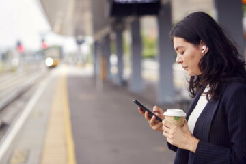 Businesswoman Waiting On Train Platform With Wireless Earbuds Listens To Music On Mobile Phone