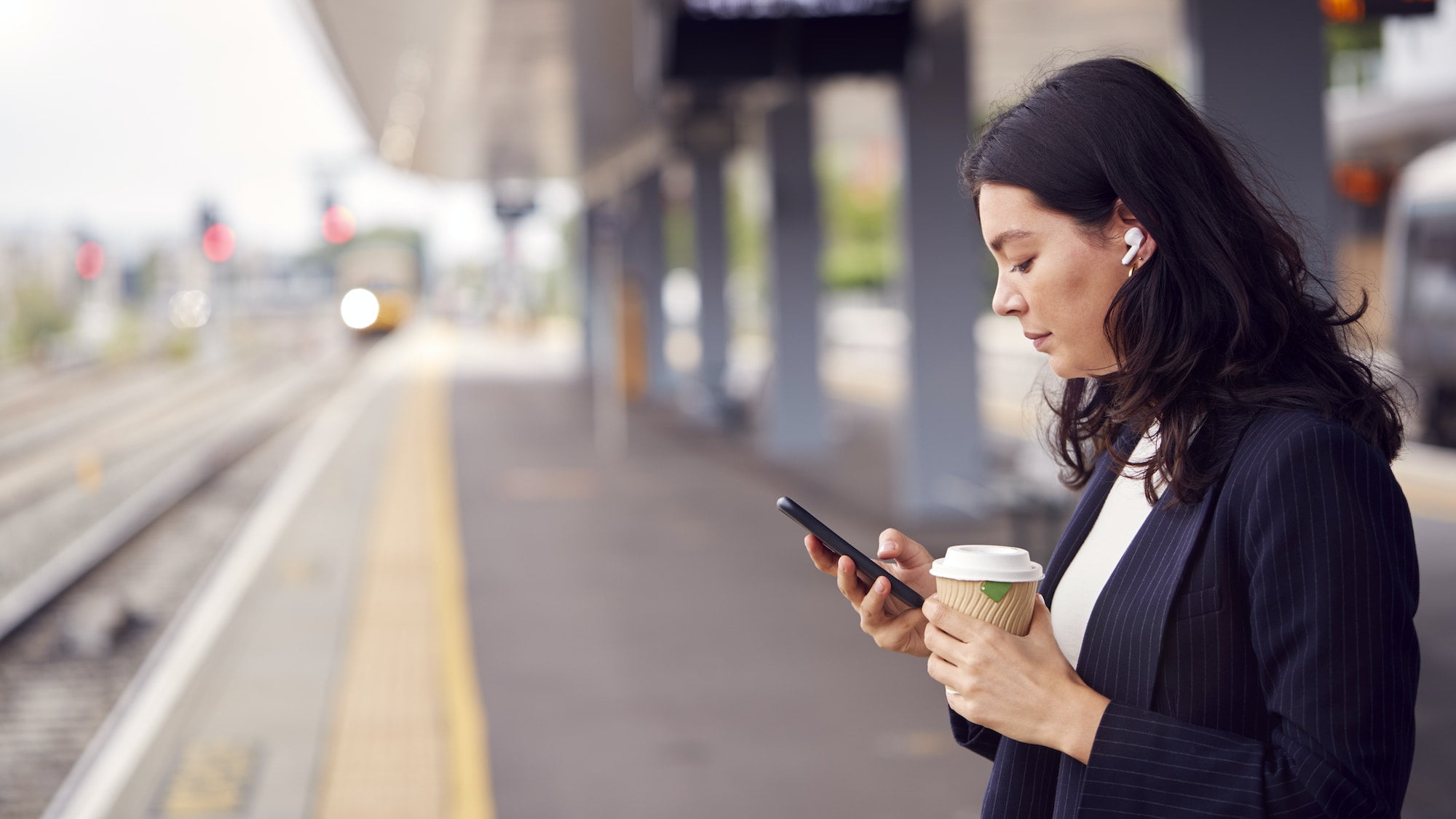 Businesswoman Waiting On Train Platform With Wireless Earbuds Listens To Music On Mobile Phone