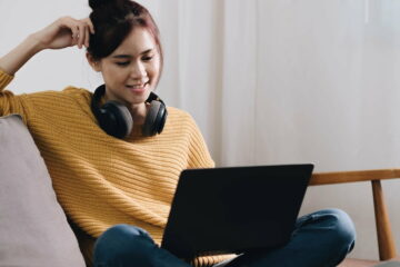 Cheerful freelancer working on laptop sitting on couch and listening to music in headphones