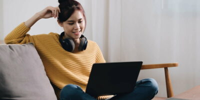 Cheerful freelancer working on laptop sitting on couch and listening to music in headphones