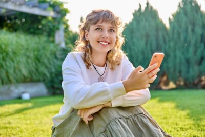 Portrait of a smiling female with smartphone in hands, sitting on grass