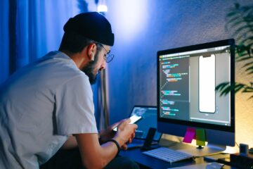 Young man mobile developer writes program code on a computer and check app in smartphone