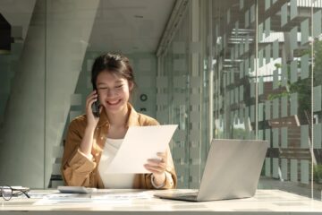 Charming Asian woman with a smile standing holding papers and mobile phone at the office.