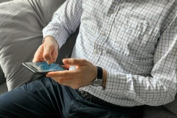 Man sitting on sofa at home before going to office and using smartphone to check weather forecast