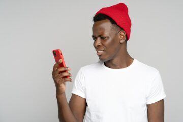 Confused African hipster man looking at mobile, annoyed by spam, intrusive adware unpleasant message