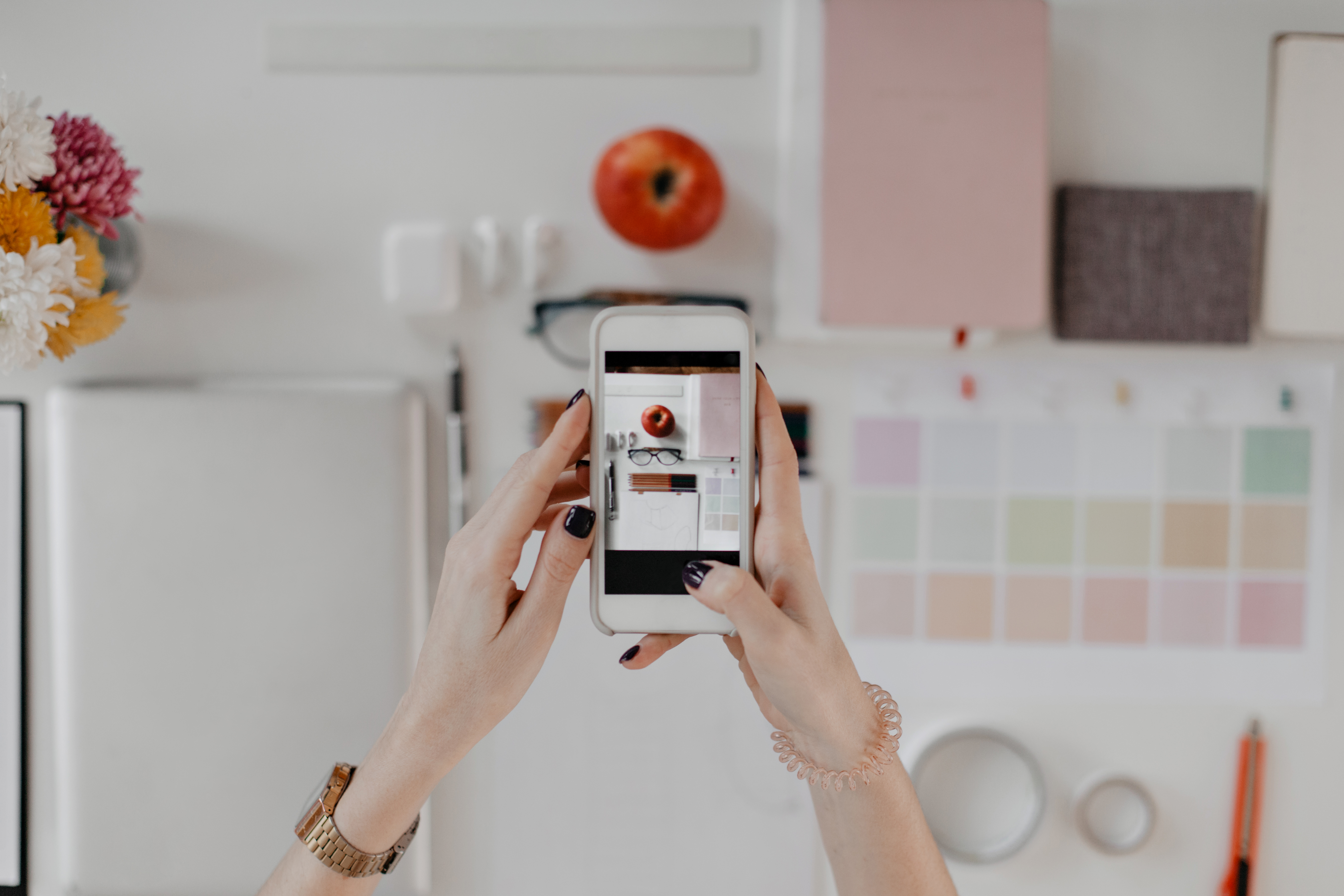 Picture of female hands taking photos of desktop with stationery, glasses and apple on iPhone