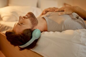 Happy man, streaming a podcast in bed smiling with headphones at home. Young guy listening to music