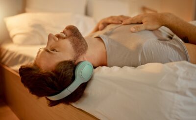 Happy man, streaming a podcast in bed smiling with headphones at home. Young guy listening to music